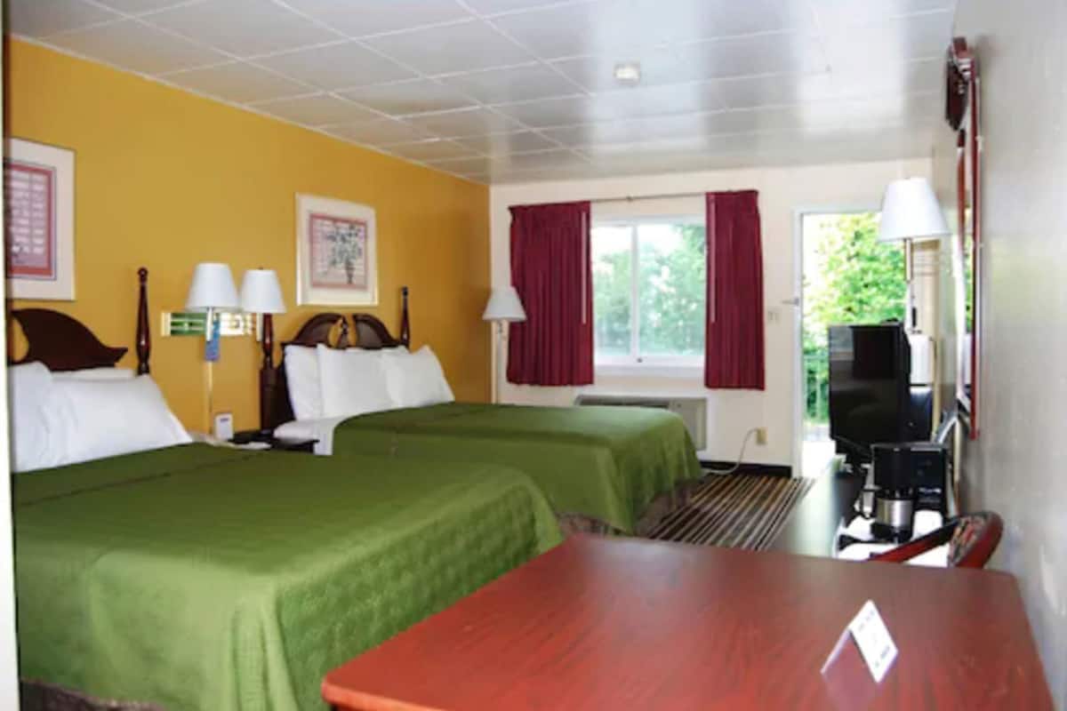 Une chambre d’hôtel avec deux lits, une table en bois, un téléviseur et une fenêtre avec des rideaux.
