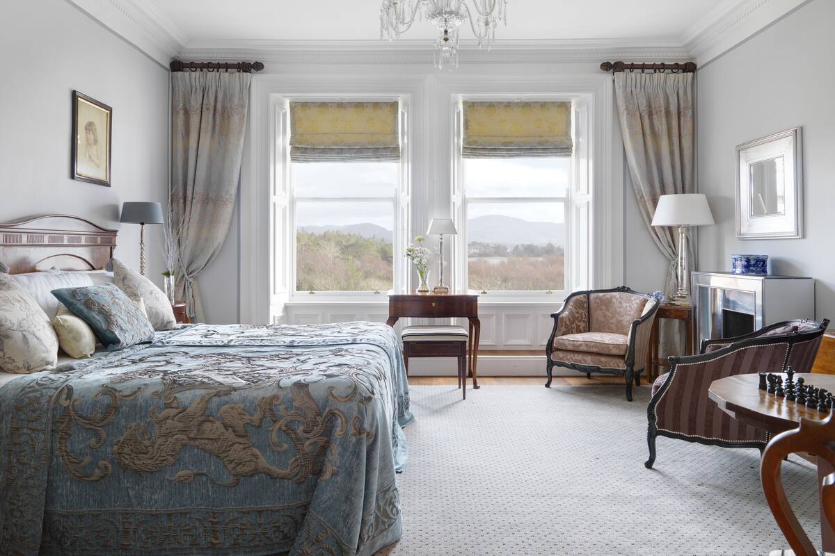 A bedroom with a large bed, two windows with curtains, a chandelier, a desk with a lamp, a chair, and a chess table.