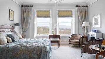 Een slaapkamer met een groot bed, twee ramen met gordijnen, een kroonluchter, een bureau met lamp, een stoel en een schaakspel.