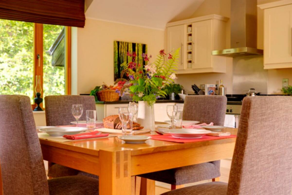 A dining area with a wooden table set for four, surrounded by chairs. There is a vase with flowers on the table, and a window with blinds to the left.