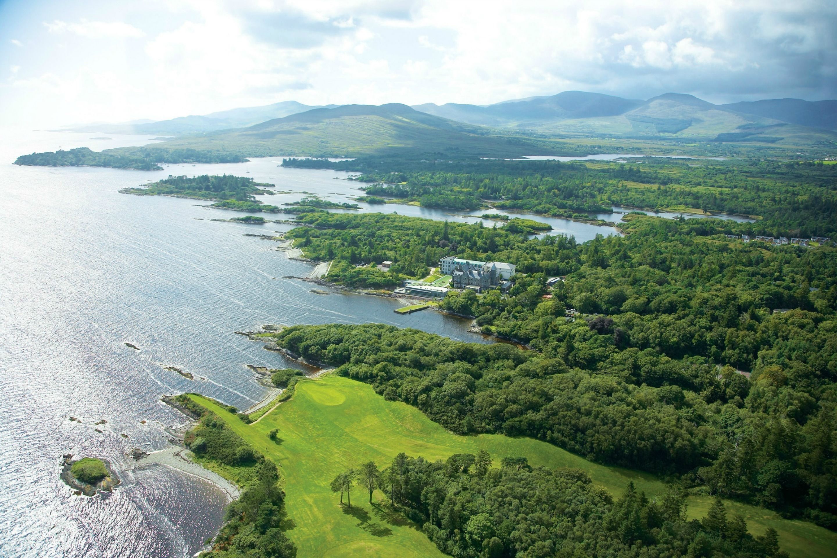 An aerial view of a coastal area with a large body of water, green hills, and a building with multiple structures.
