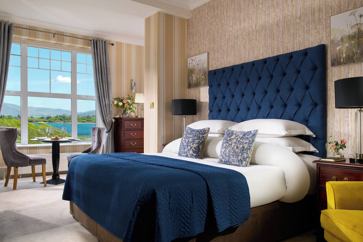 A hotel room with a large bed, a view of the outdoors, a desk with a chair, and a nightstand with a lamp.