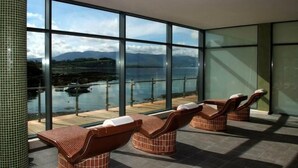 A spa area with three wooden loungers and a large glass window offering a view of a lake and mountains. - Parknasilla Resort and Spa (Sneem)