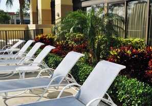 Een rij witte lounge stoelen met metalen frames, geplaatst in een zonnig buitengebied met verschillende groene struiken en rode bloemen op de achtergrond.