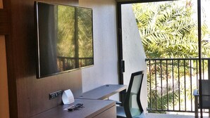 A hotel room with a large flat-screen TV mounted on the wall, a wooden desk with a chair, and a balcony with a view of palm trees. - Holiday Inn Express Boca Raton-West by IHG (Boca Raton)