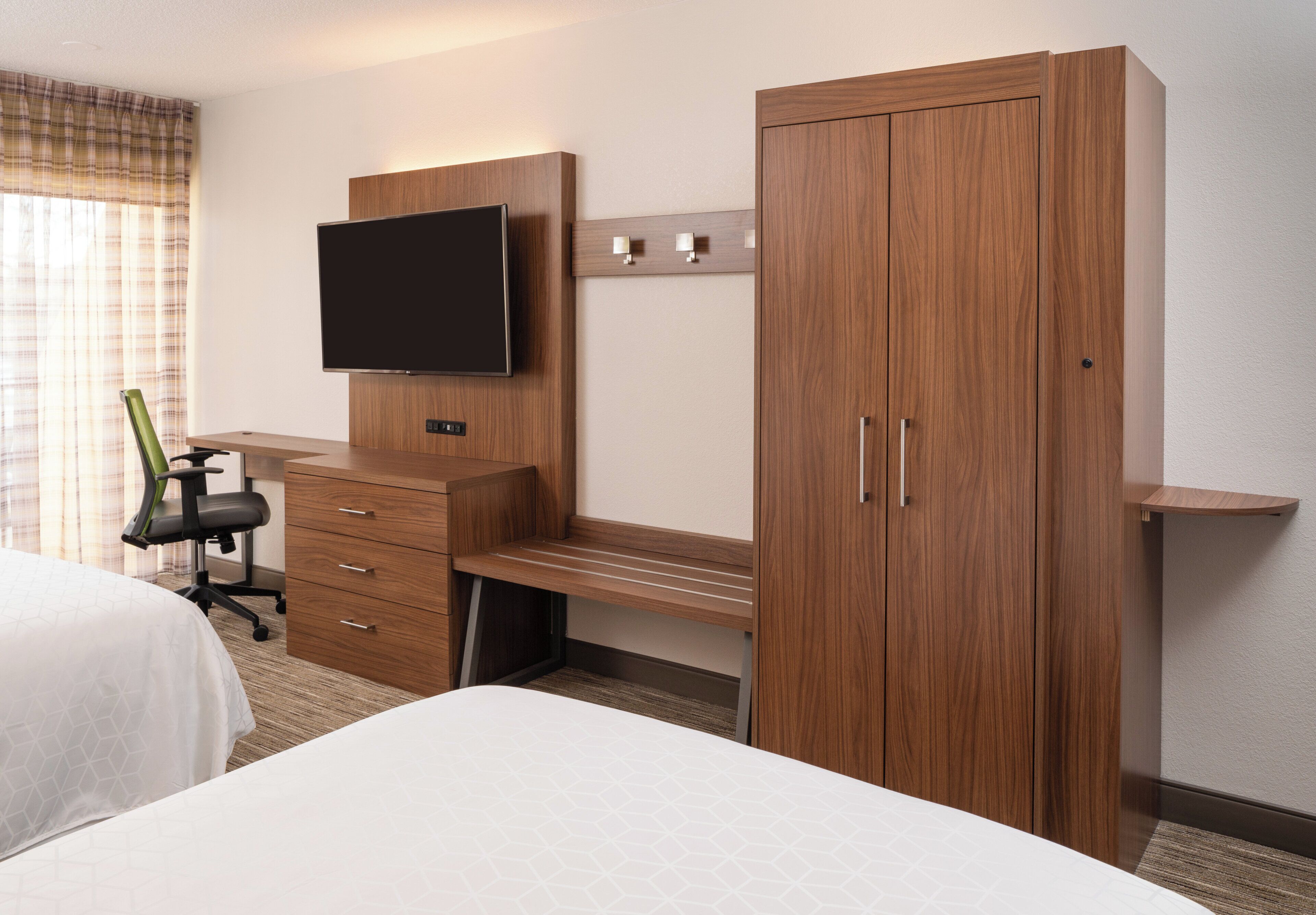 A hotel room with a bed, a desk with a chair, a television mounted on the wall, and a wardrobe.