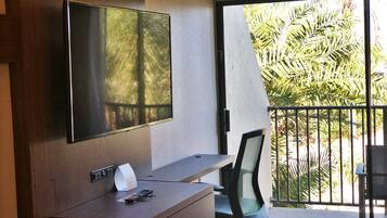 A hotel room with a flat-screen TV mounted on the wall, a desk with a chair, and a balcony with a view of palm trees.
