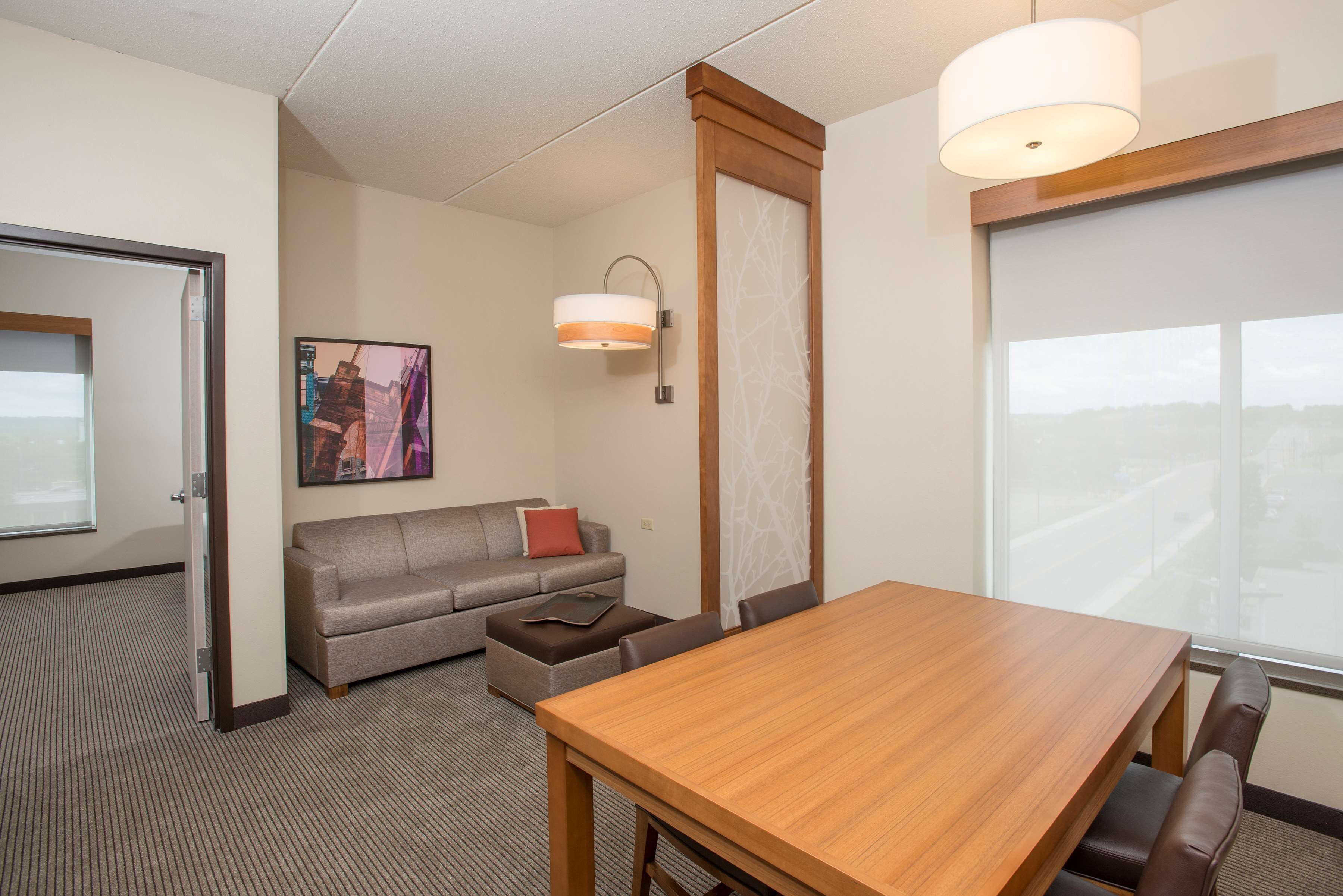 Une chambre d’hôtel avec une table à manger en bois, un canapé gris agrémenté d’un coussin rouge, un petit pouf, une lampe fixée au mur et une grande fenêtre donnant sur la ville.