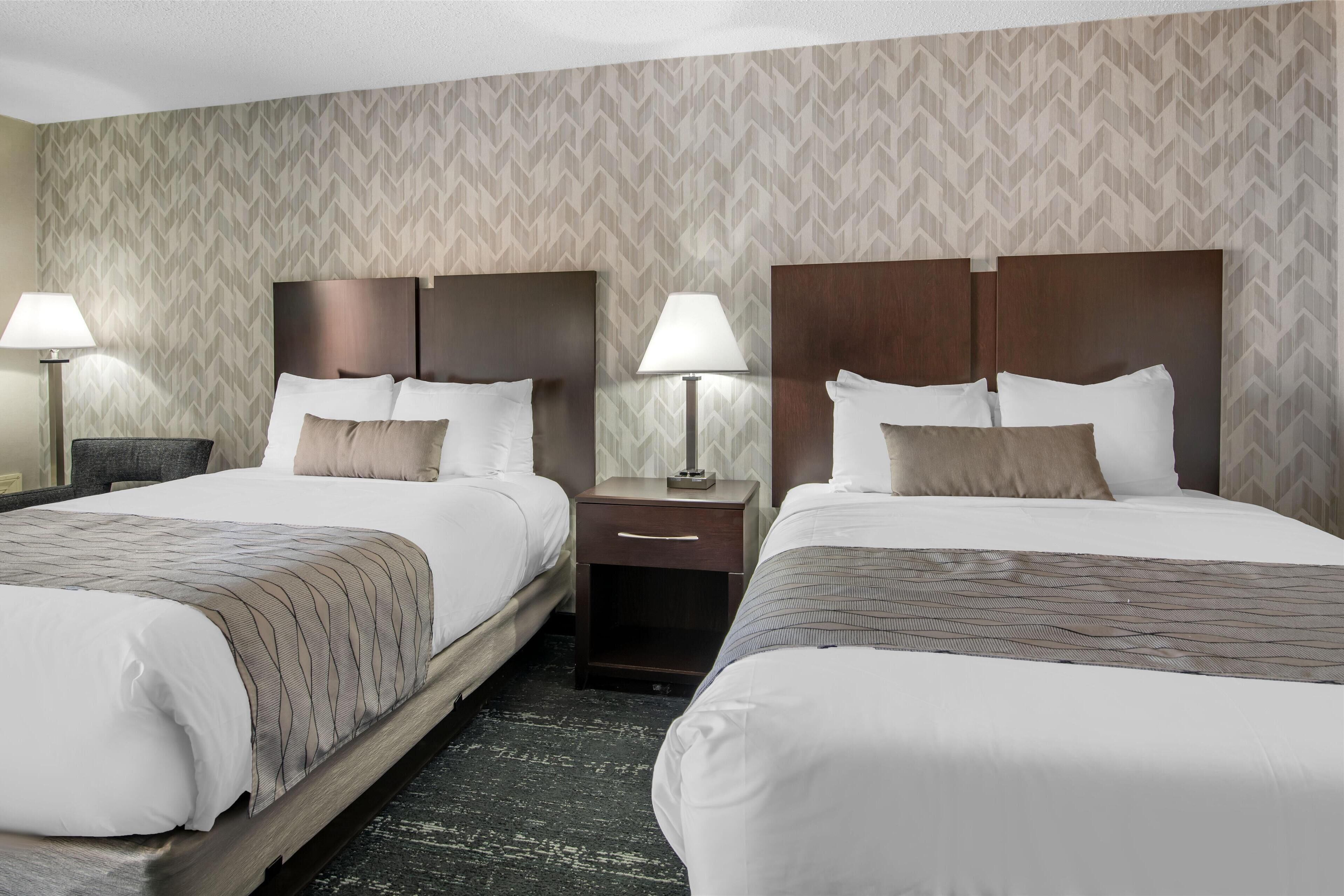 A hotel room with two beds, a nightstand with a lamp, and a patterned wallpaper.