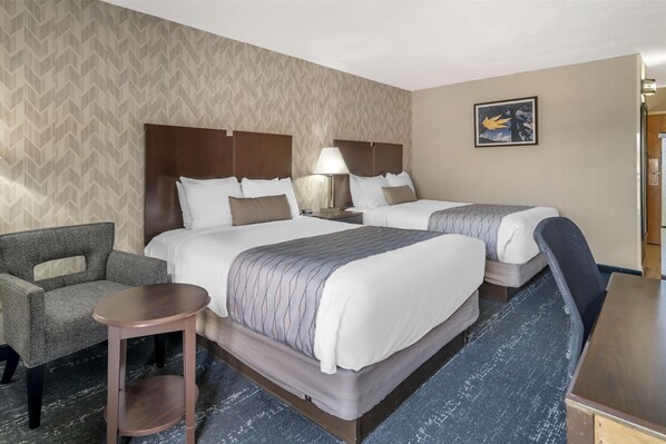 A hotel room with two beds, a chair, a small table, and a lamp.