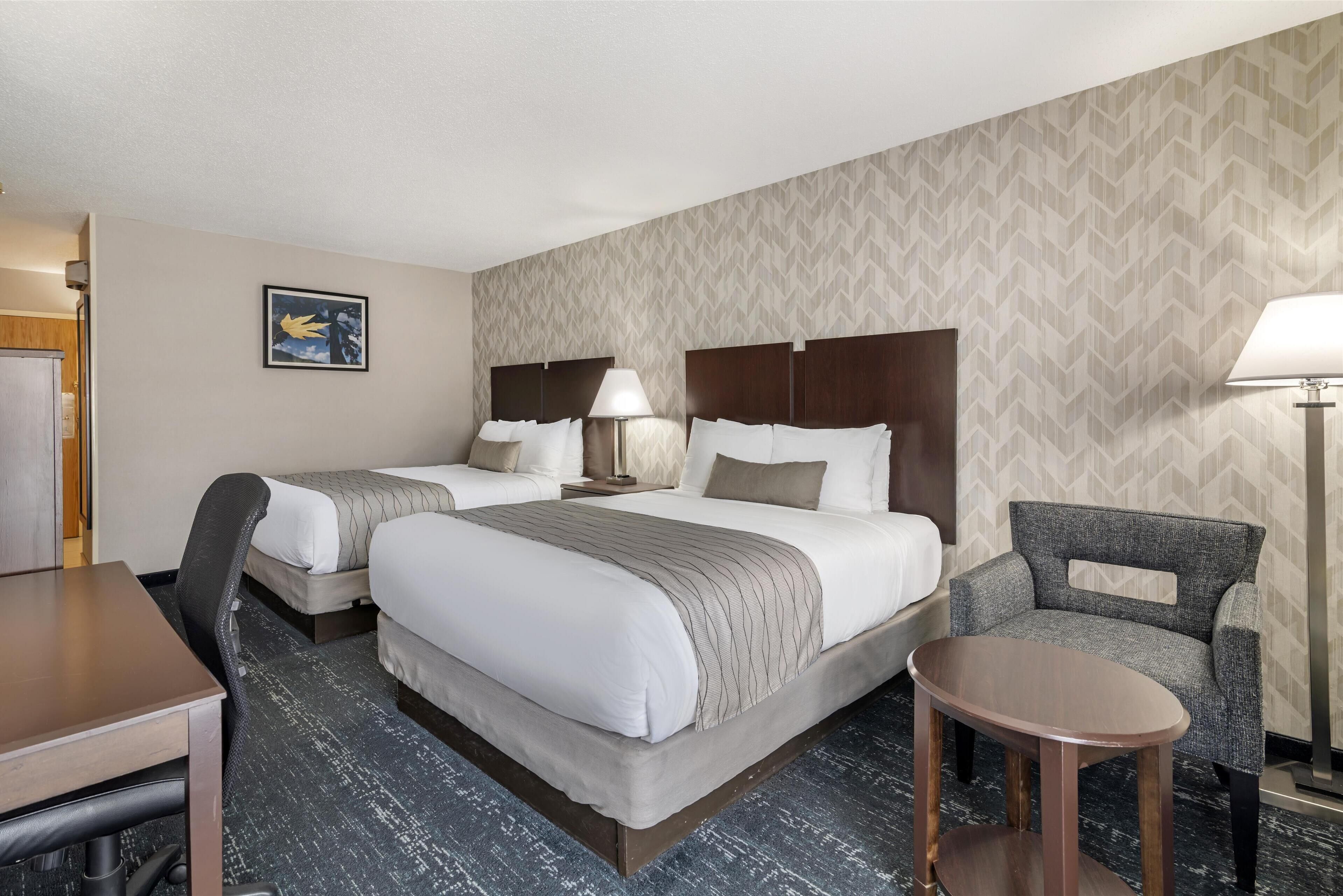 Quarto de hotel com duas camas, uma escrivaninha, uma cadeira e uma mesinha.