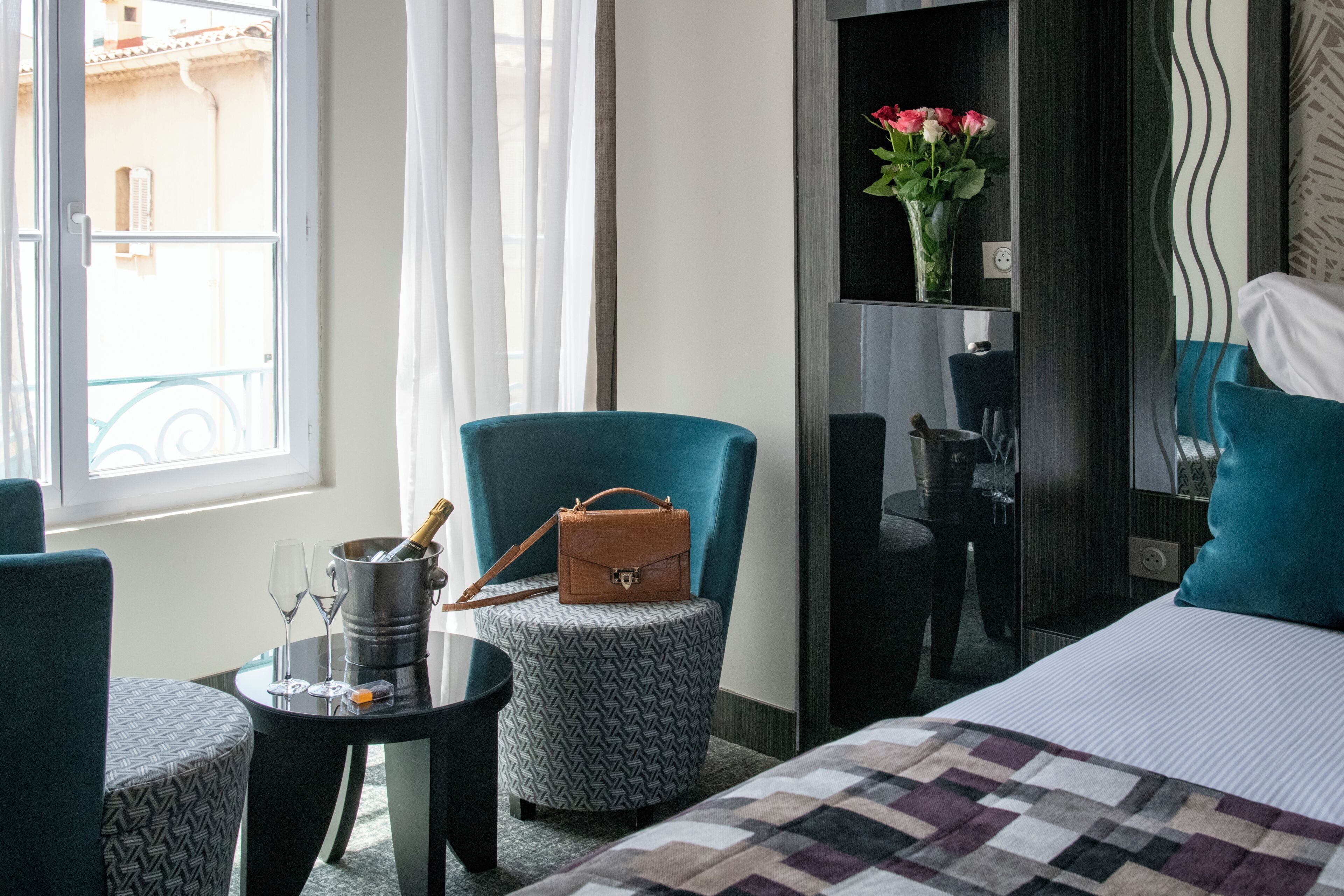 A hotel room with a bed, a small round table with a chair, a minibar, a minibar shelf with a vase of flowers, and a window with curtains.