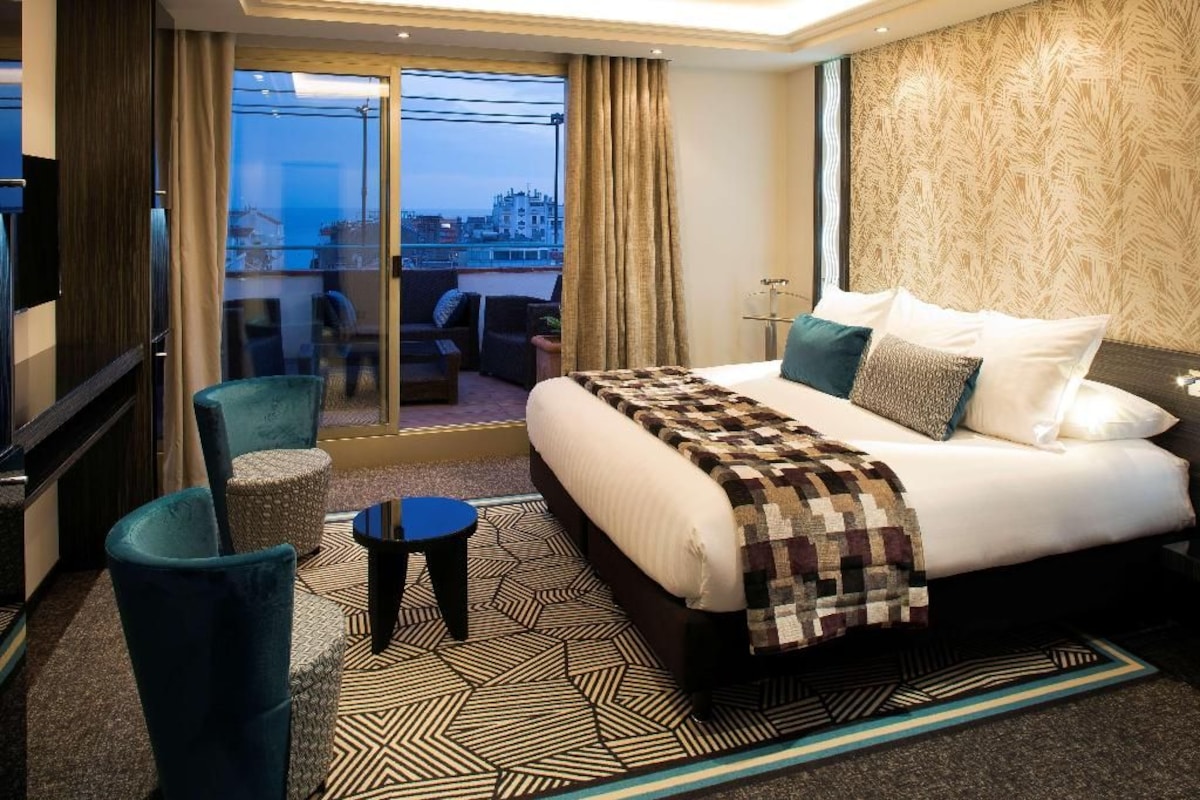 Habitación de hotel con una cama grande, una zona de estar con sillas, una mesita y vistas a la ciudad a través de un ventanal.