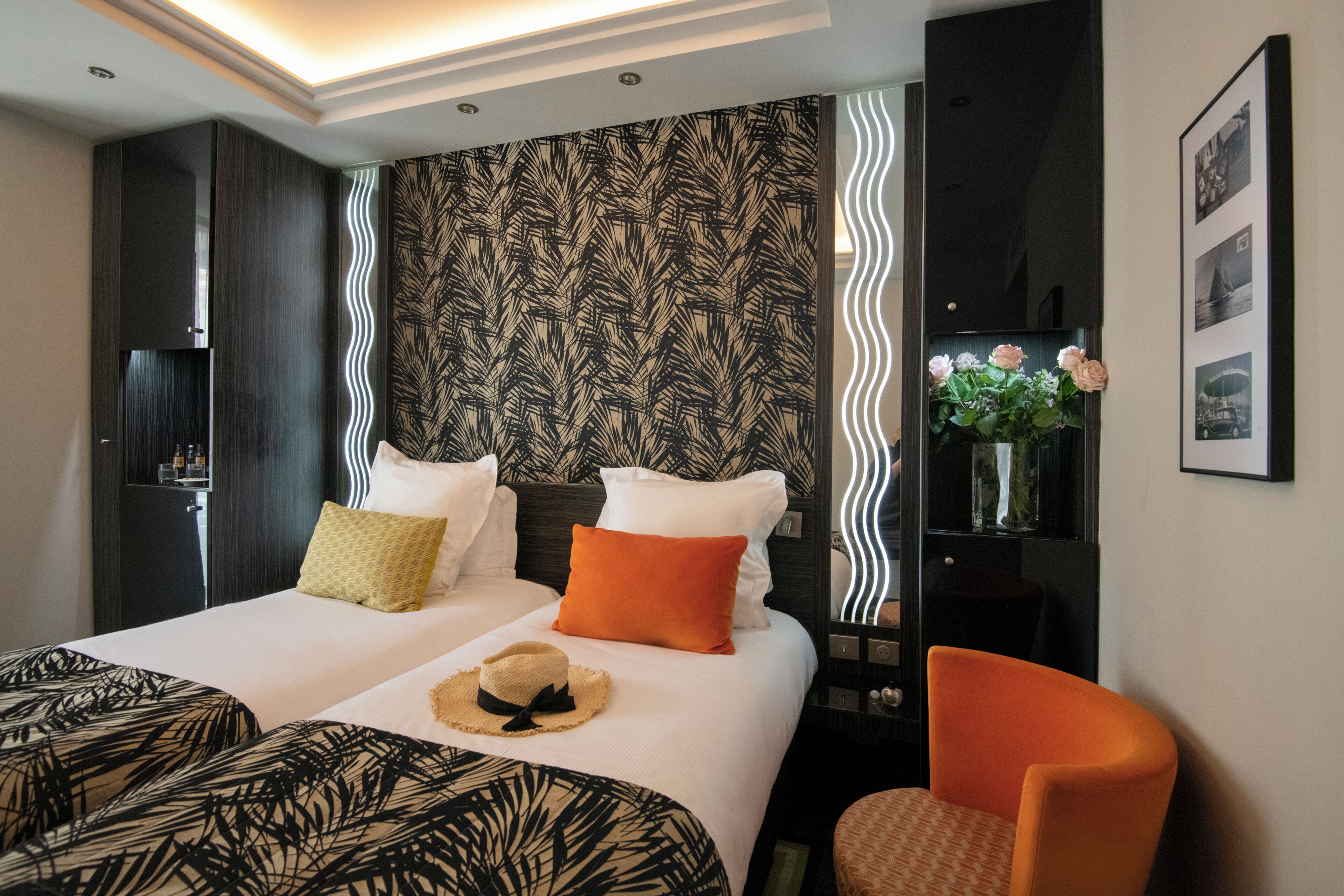 Une chambre d’hôtel avec deux lits, une table de chevet, une chaise, un miroir et un mur orné d’un motif de feuilles.