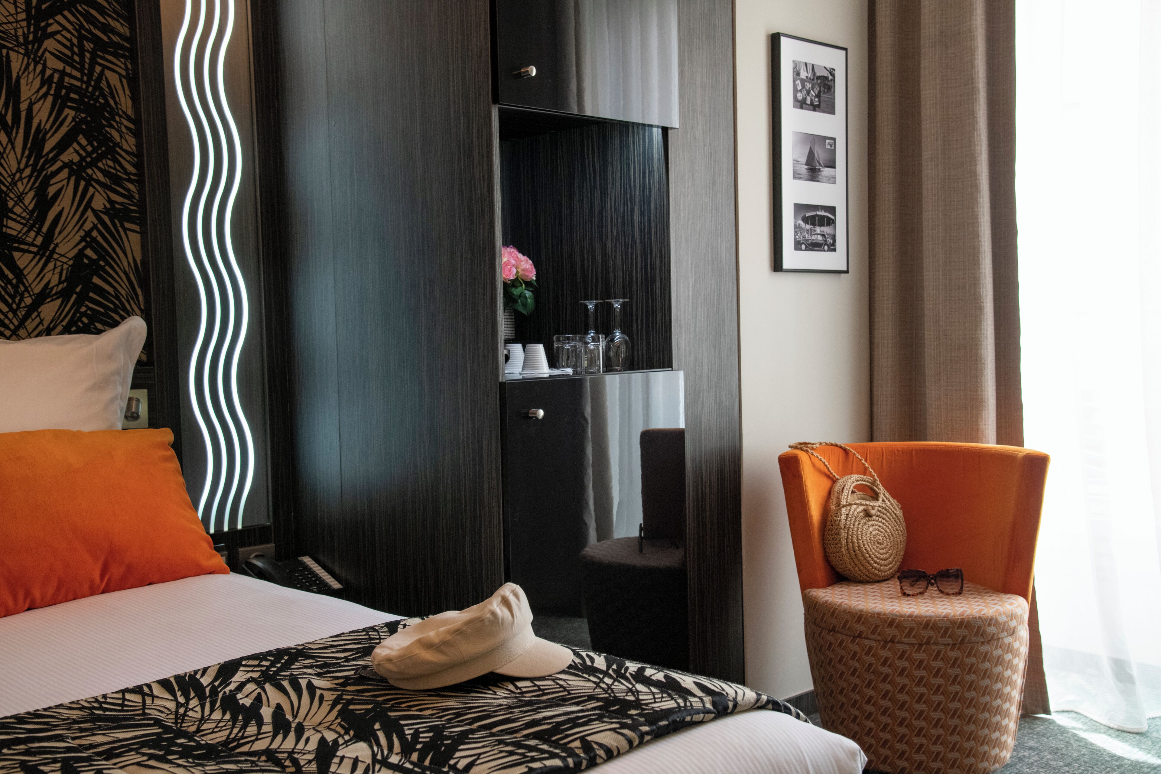 A hotel room with a bed, a chair, a wardrobe, and a shelf with glasses and a handbag.
