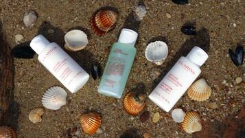 Three bottles of Clarins body lotion on a sandy surface with seashells.