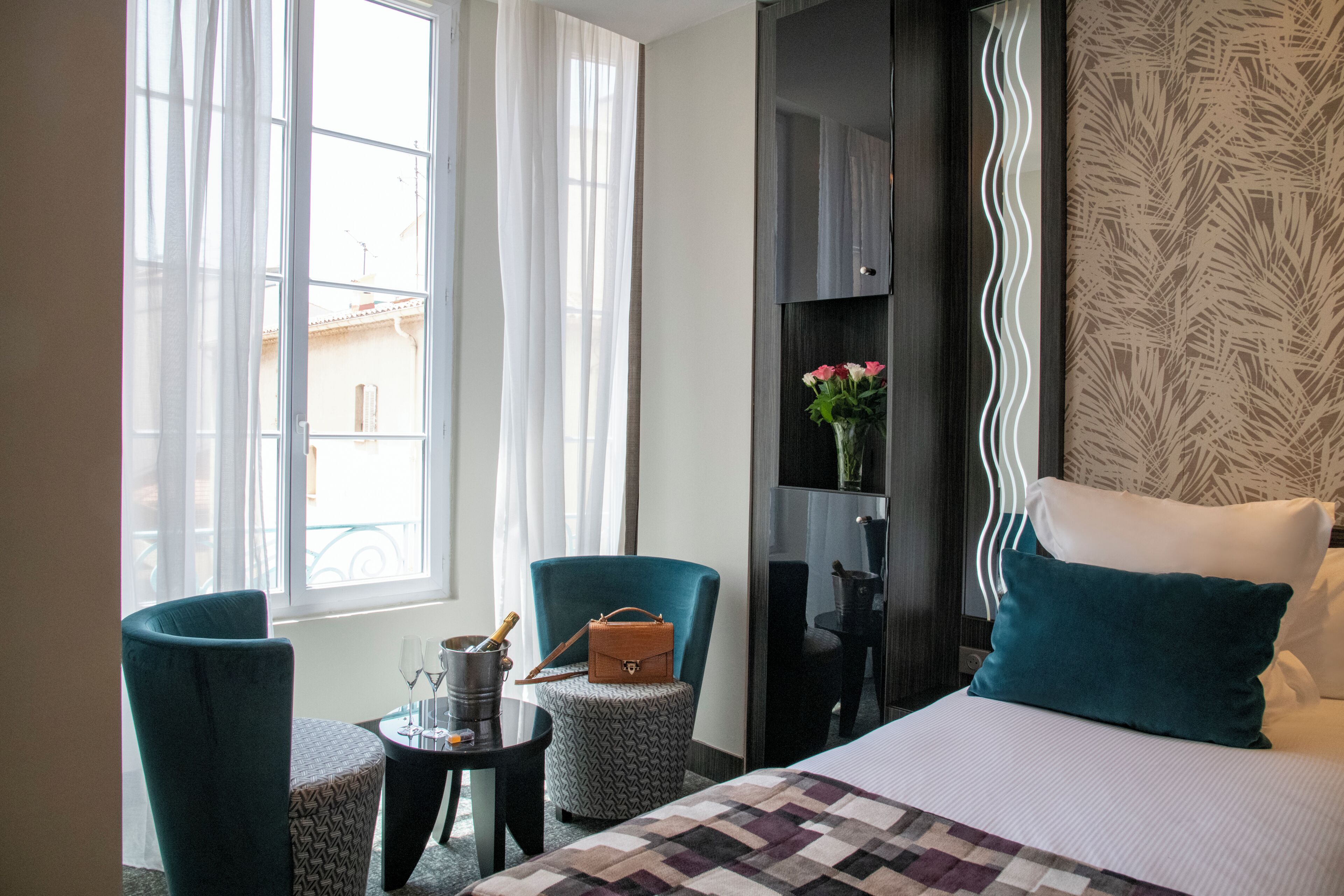 A hotel room with a bed, two chairs, a small table with a bottle and glasses, and a vase with flowers.