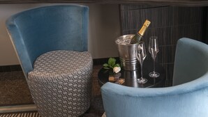A room with a blue upholstered chair, a patterned ottoman, a small round table with a champagne bottle, glasses, and a rose. - BW Premier Collection Mondial (Cannes)