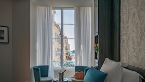 A hotel room with a bed, two chairs, a small table with a bottle and glasses, and a view of buildings through a window. - BW Premier Collection Mondial (Cannes)