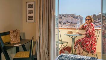 A room with a bed, a chair, a table, and a view of the cityscape through a window.