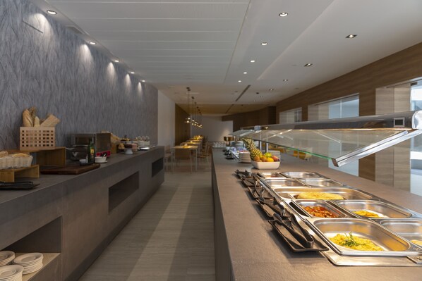 Zona de buffet con varias estaciones de comida, un mostrador con pan y acompañamientos, y un área de estar al fondo.