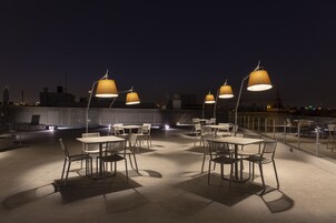 An outdoor dining area with tables and chairs, illuminated by overhead lamps, set against a cityscape at night.