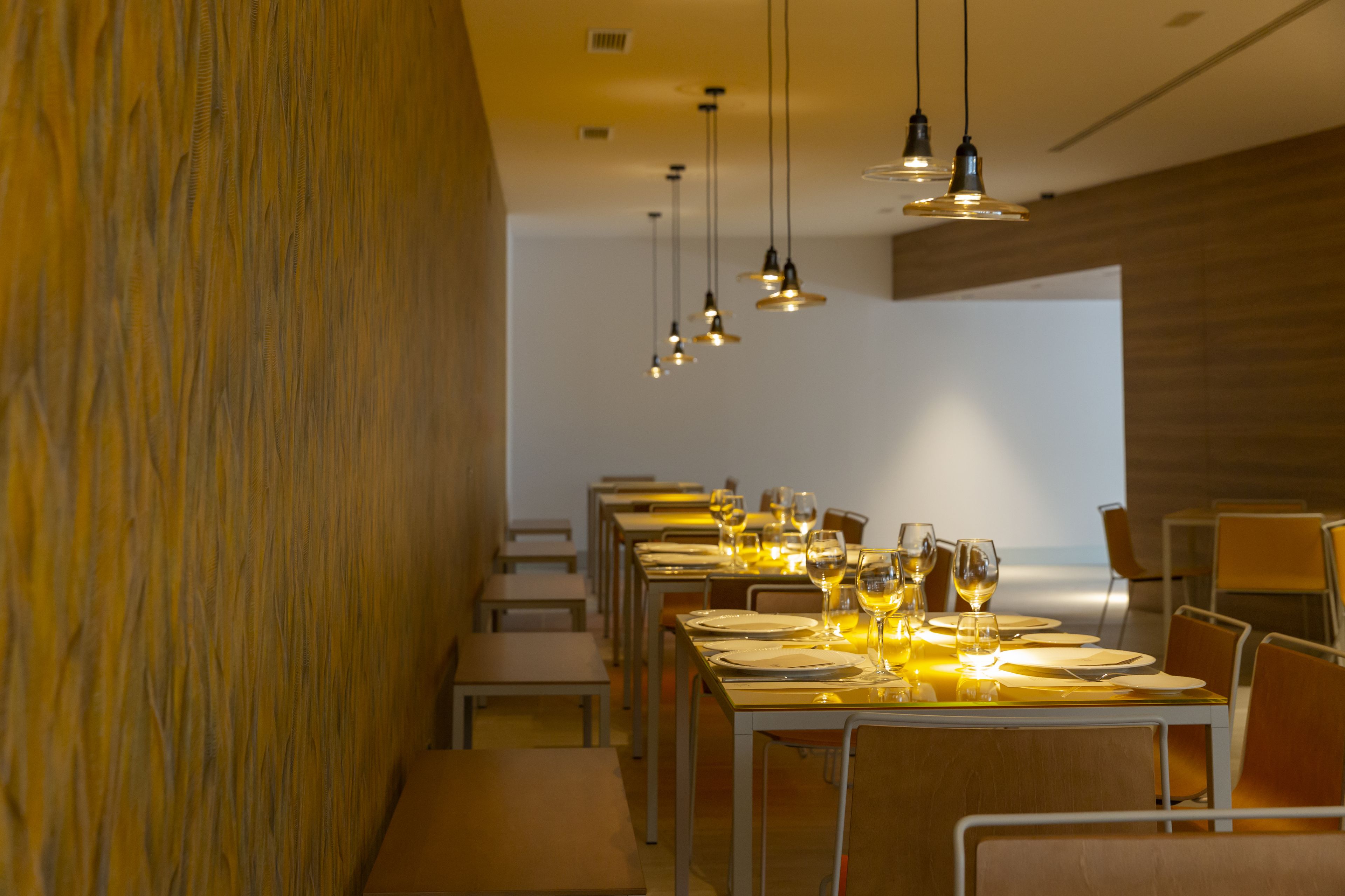 Un restaurante con largas mesas dispuestas para comer, iluminación colgante y vista al océano a través de un amplio ventanal.