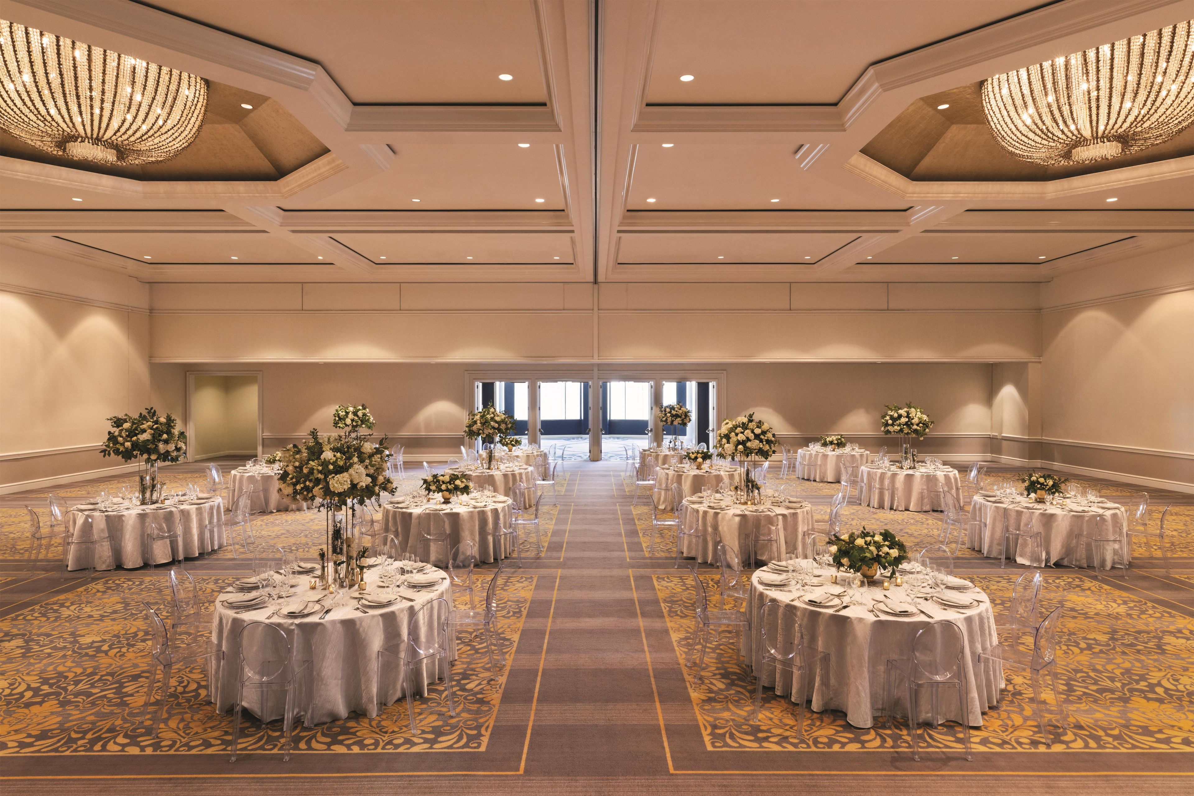 A large, well-lit banquet hall with round tables, clear chairs, and floral centerpieces.
