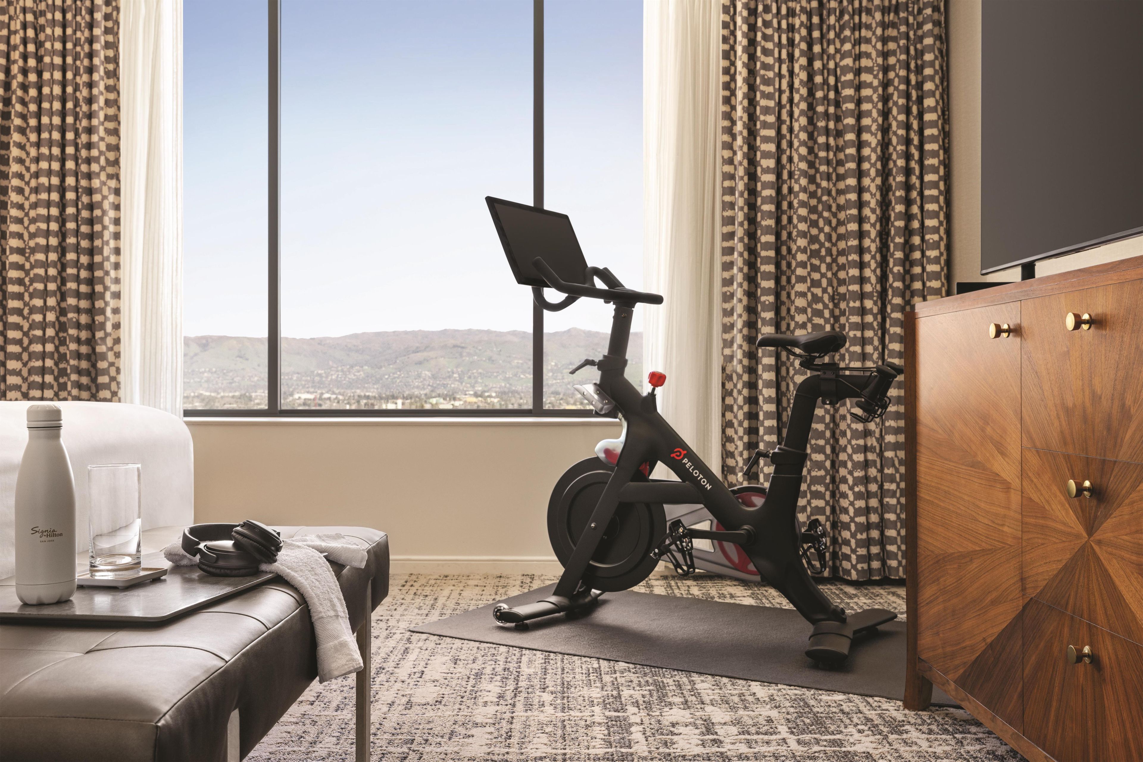 Habitación con vistas a un paisaje urbano, un escritorio con una botella de agua, un vaso y un par de auriculares, además de una bicicleta estática.