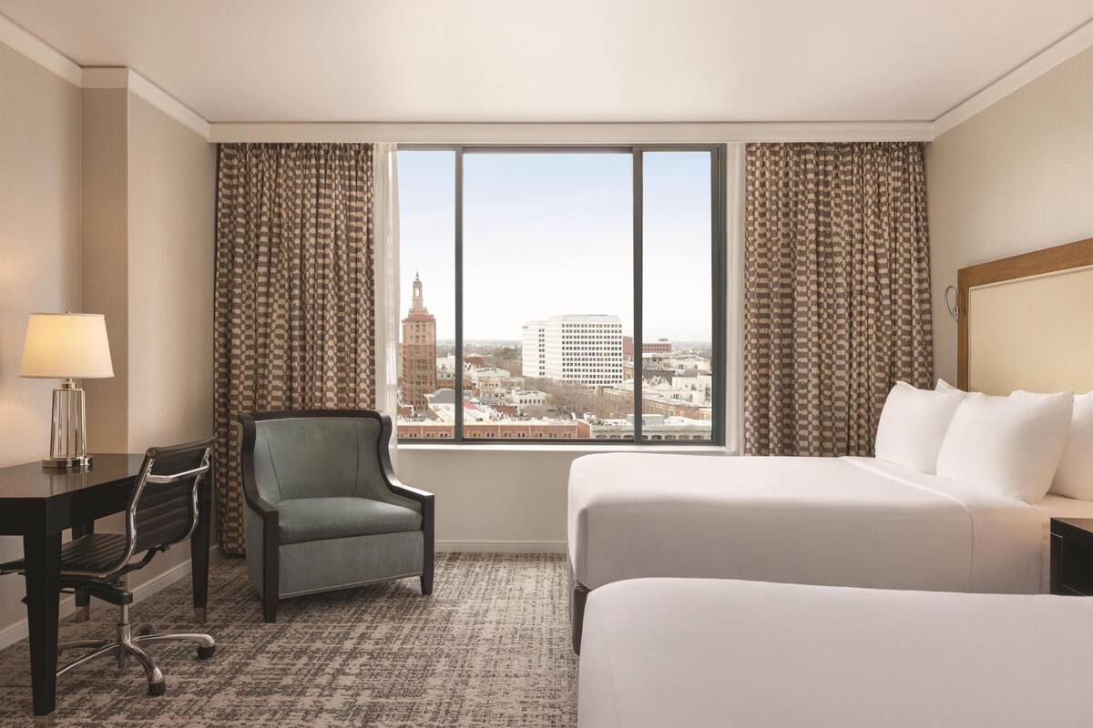 A hotel room with a large window, a desk with a lamp, a chair, and a bed.