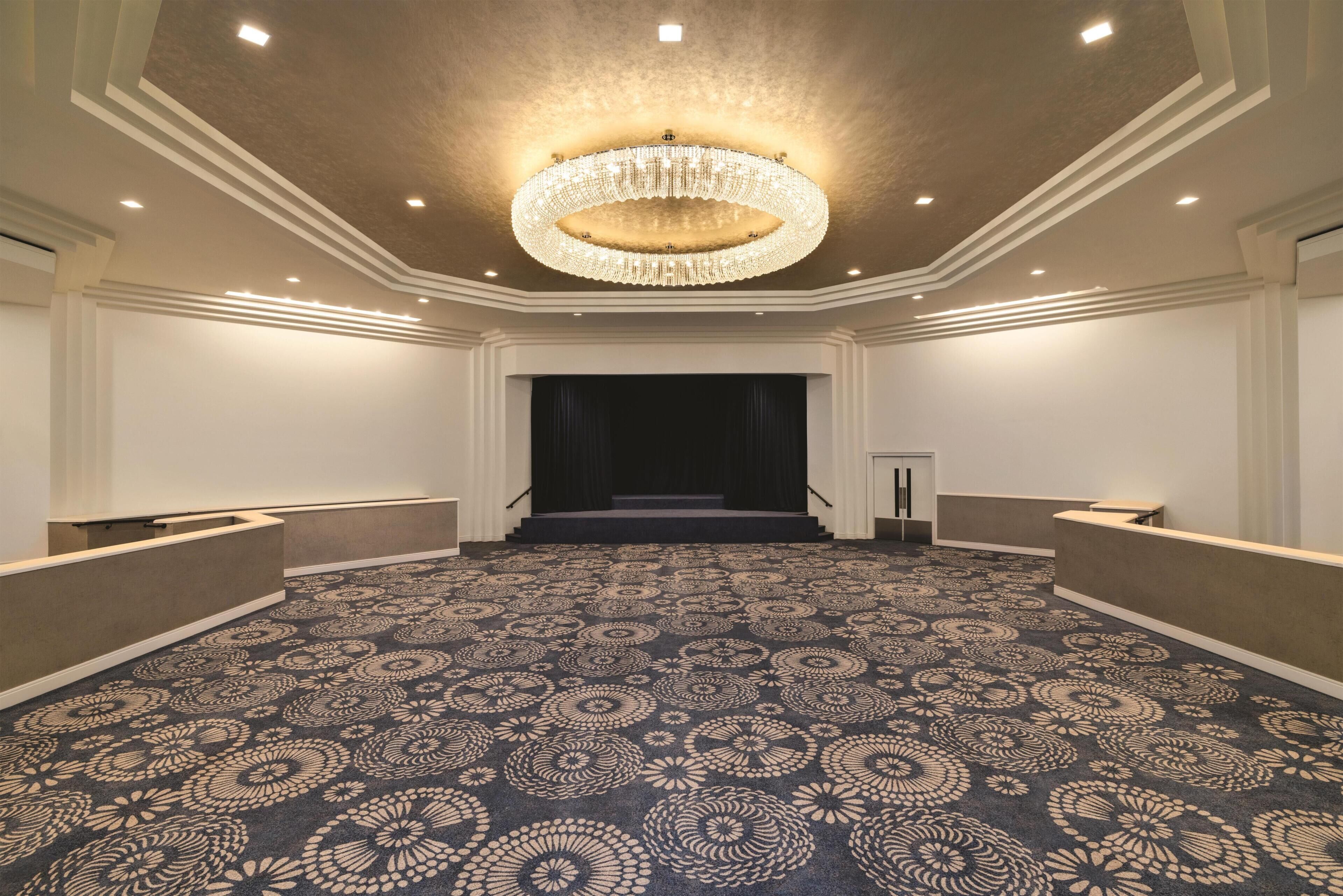 A spacious hotel lobby with a patterned carpet, a grand chandelier, and a stage at the far end.