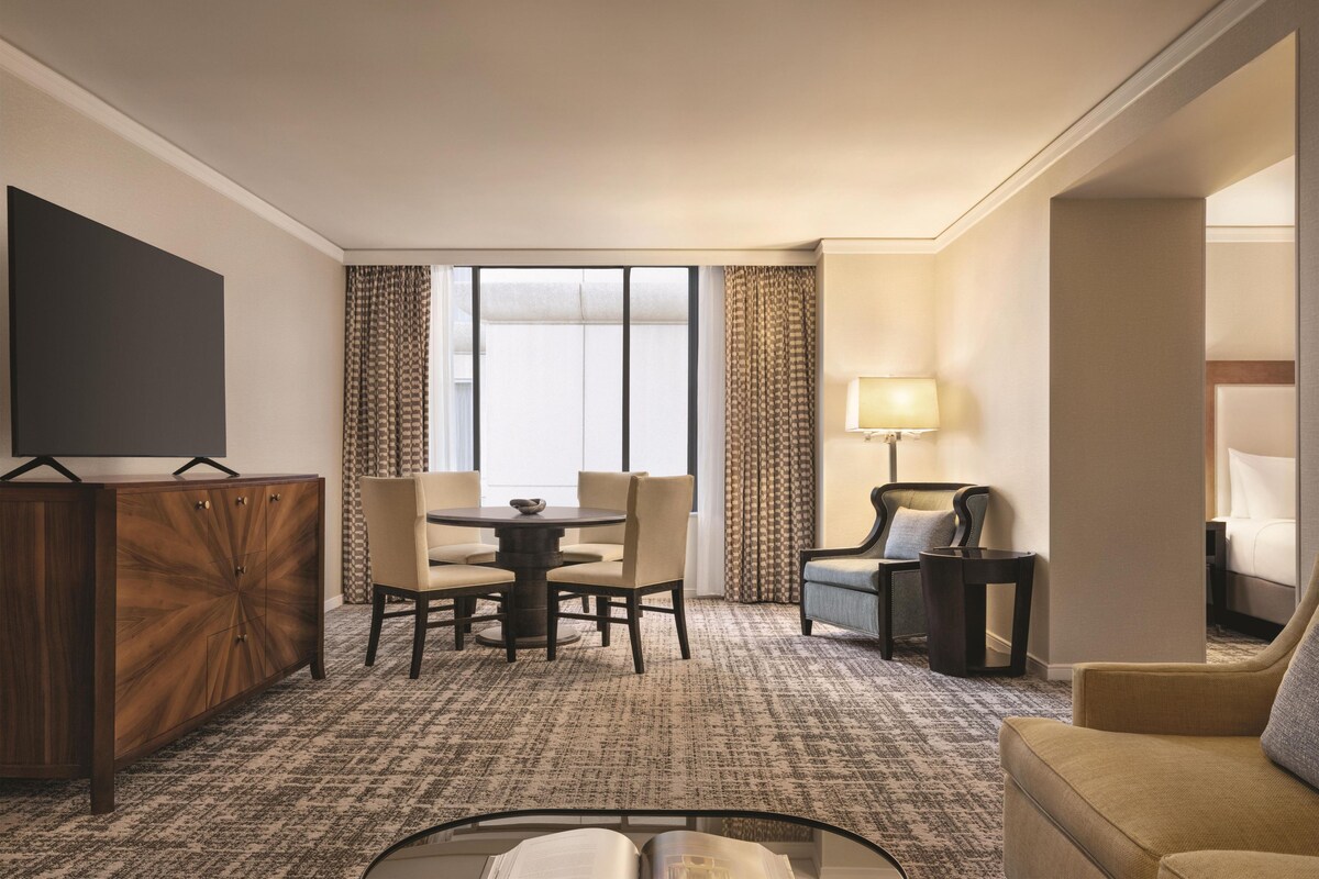 A hotel room with a dining table, chairs, a sofa, a TV, a lamp, and a large window.