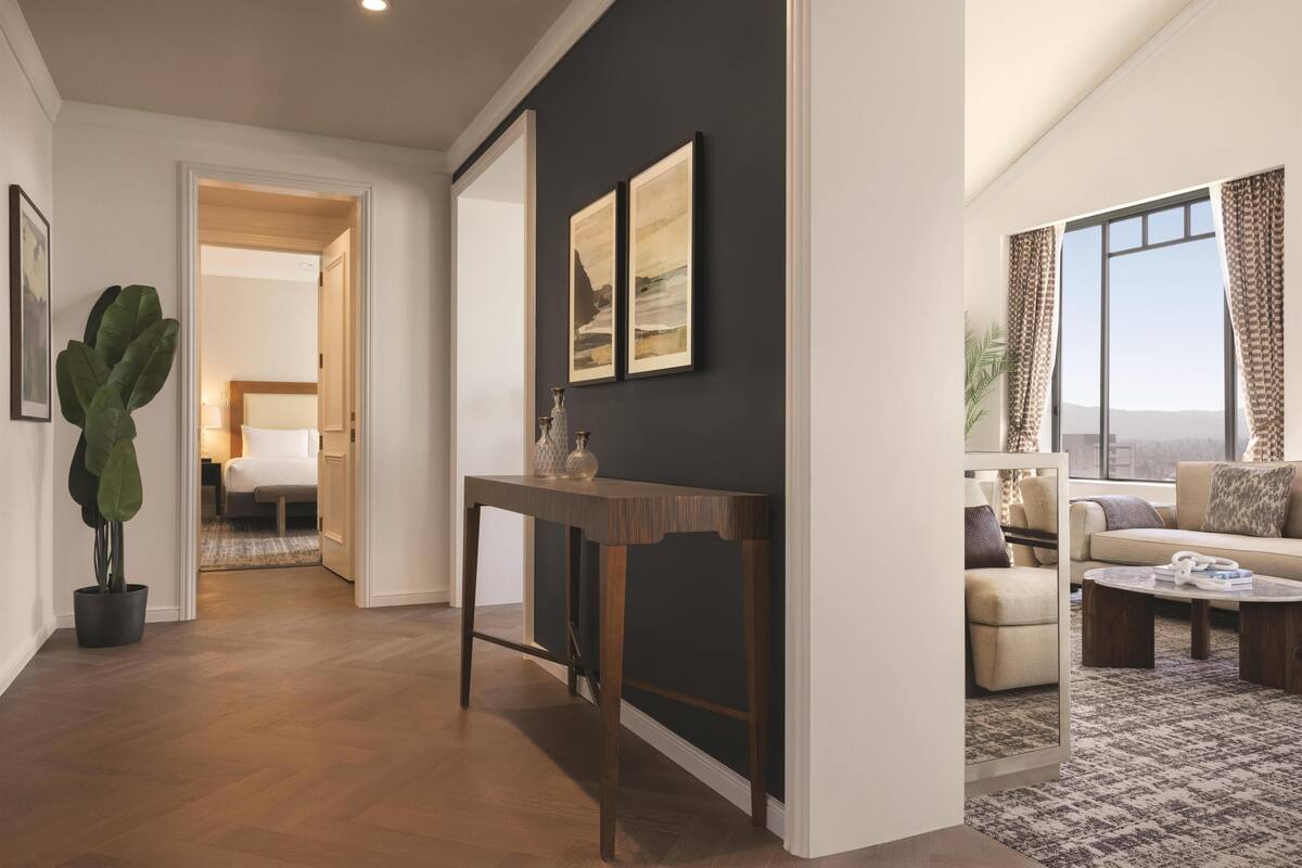A modern hotel room with a wooden floor, a dark accent wall, a dining table, a sofa, and a large window.