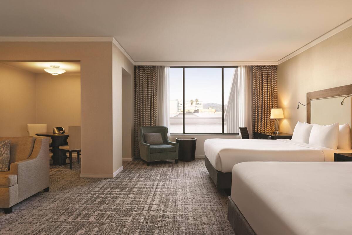 A hotel room with two beds, a chair, a small table, and a view of the city.