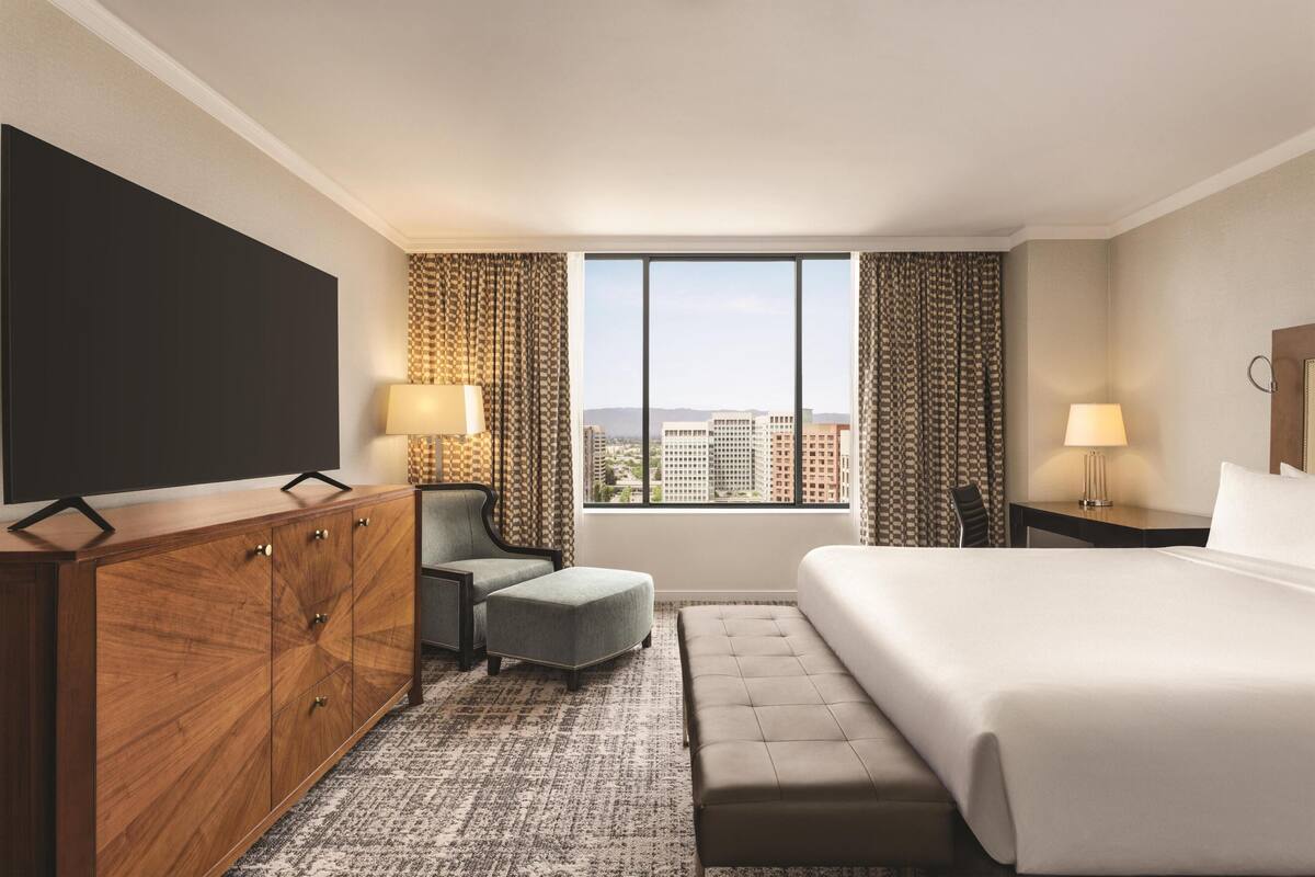A hotel room with a large bed, a TV, a desk, a chair, and a view of the city.