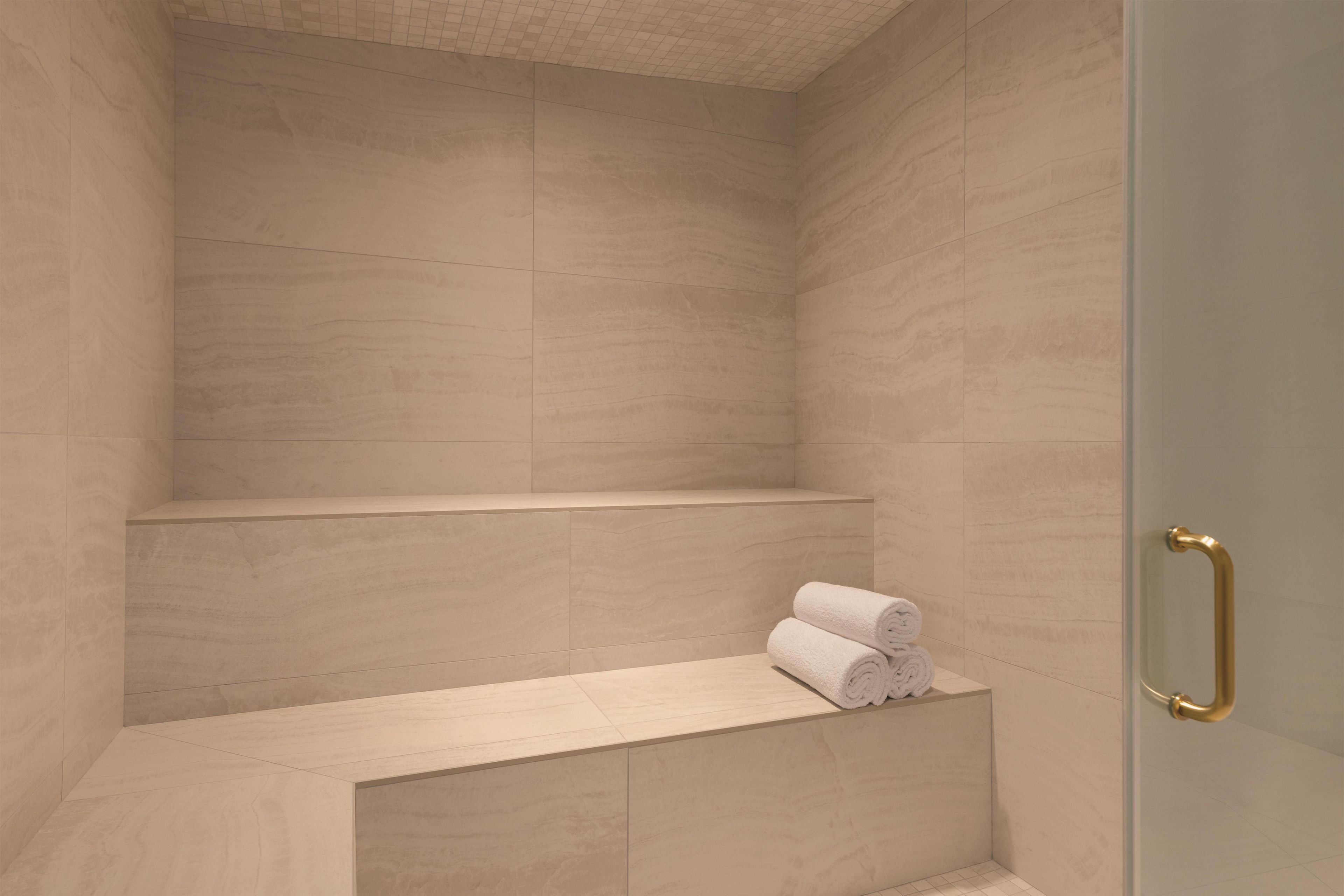 A modern bathroom with a shower area, a bench with towels, and a glass door with a gold handle.