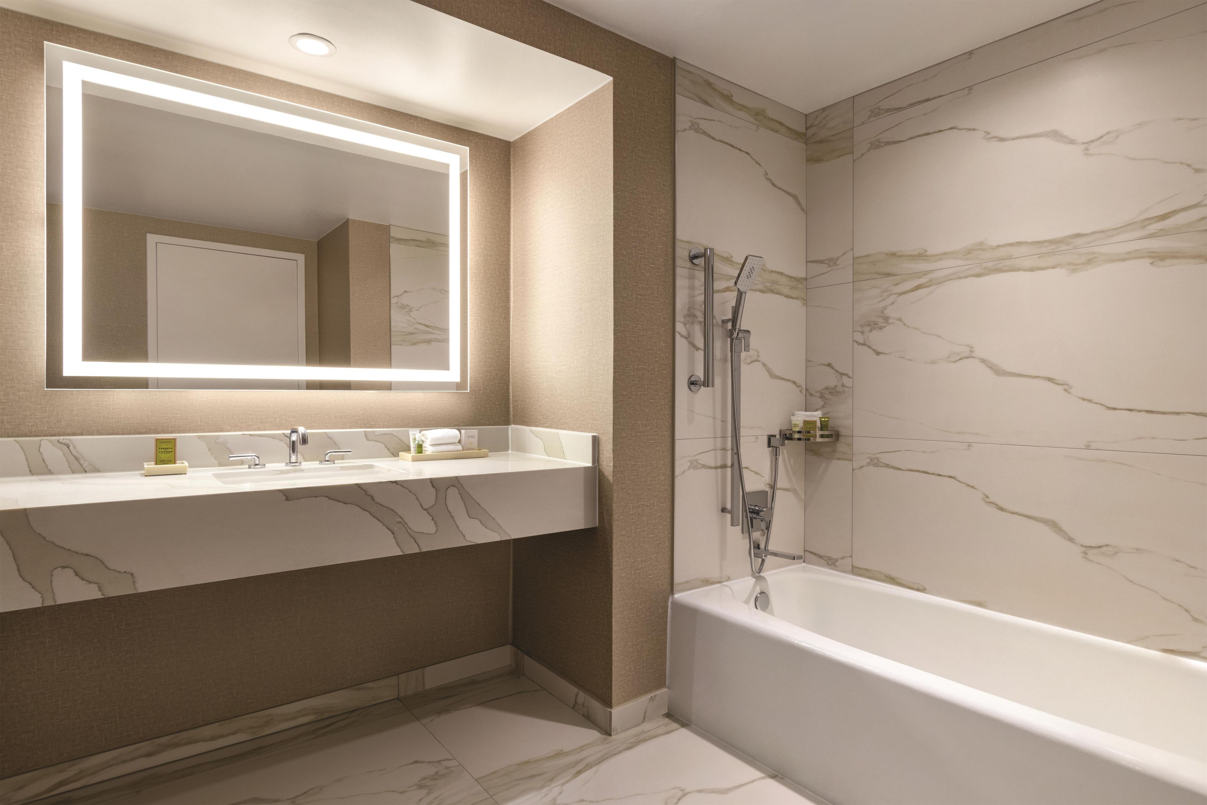 A bathroom with a marble vanity, a large mirror with integrated lighting, a bathtub, and a shower with a handheld showerhead.