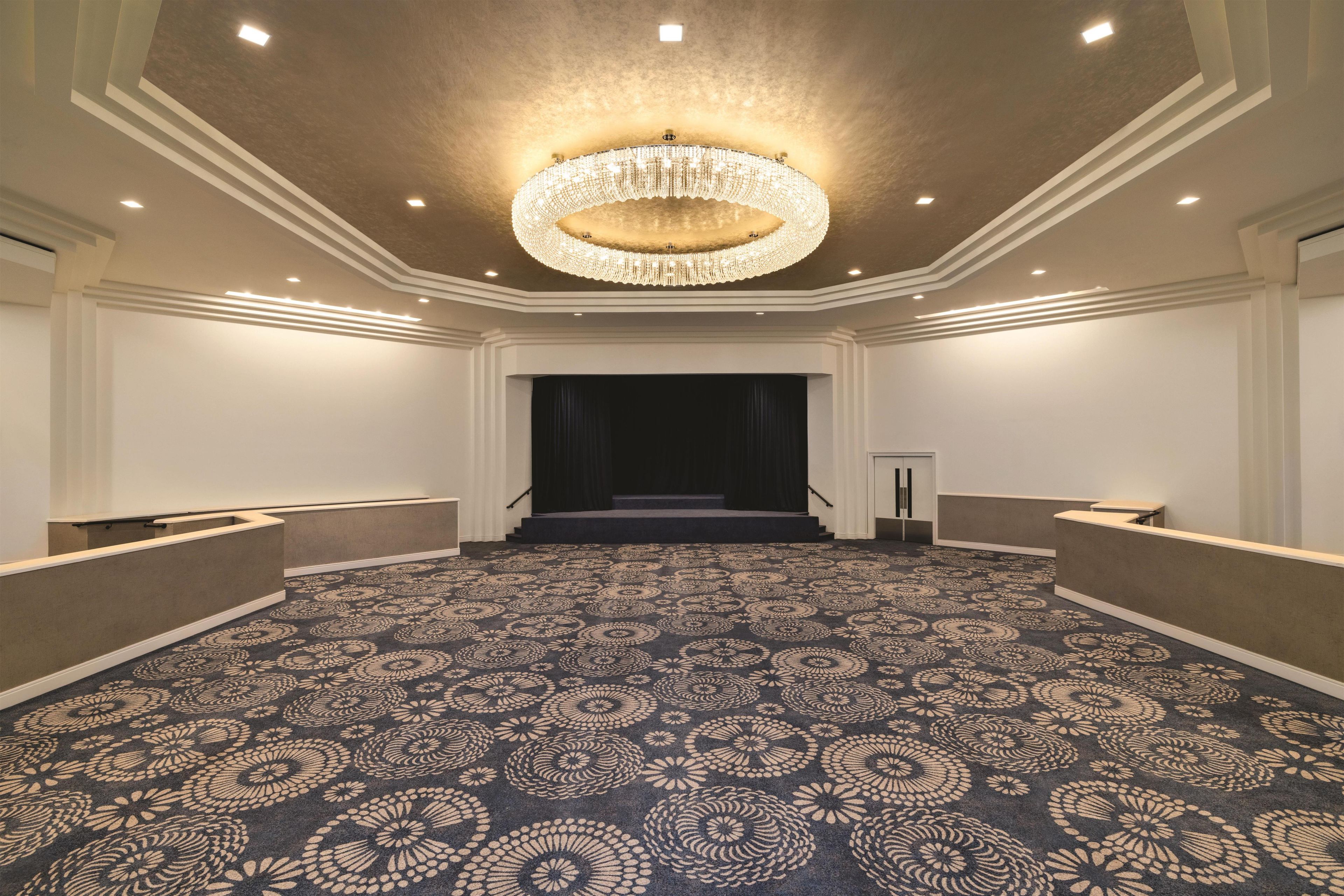 A spacious hotel lobby with a patterned carpet, a grand chandelier, and a stage at the far end.