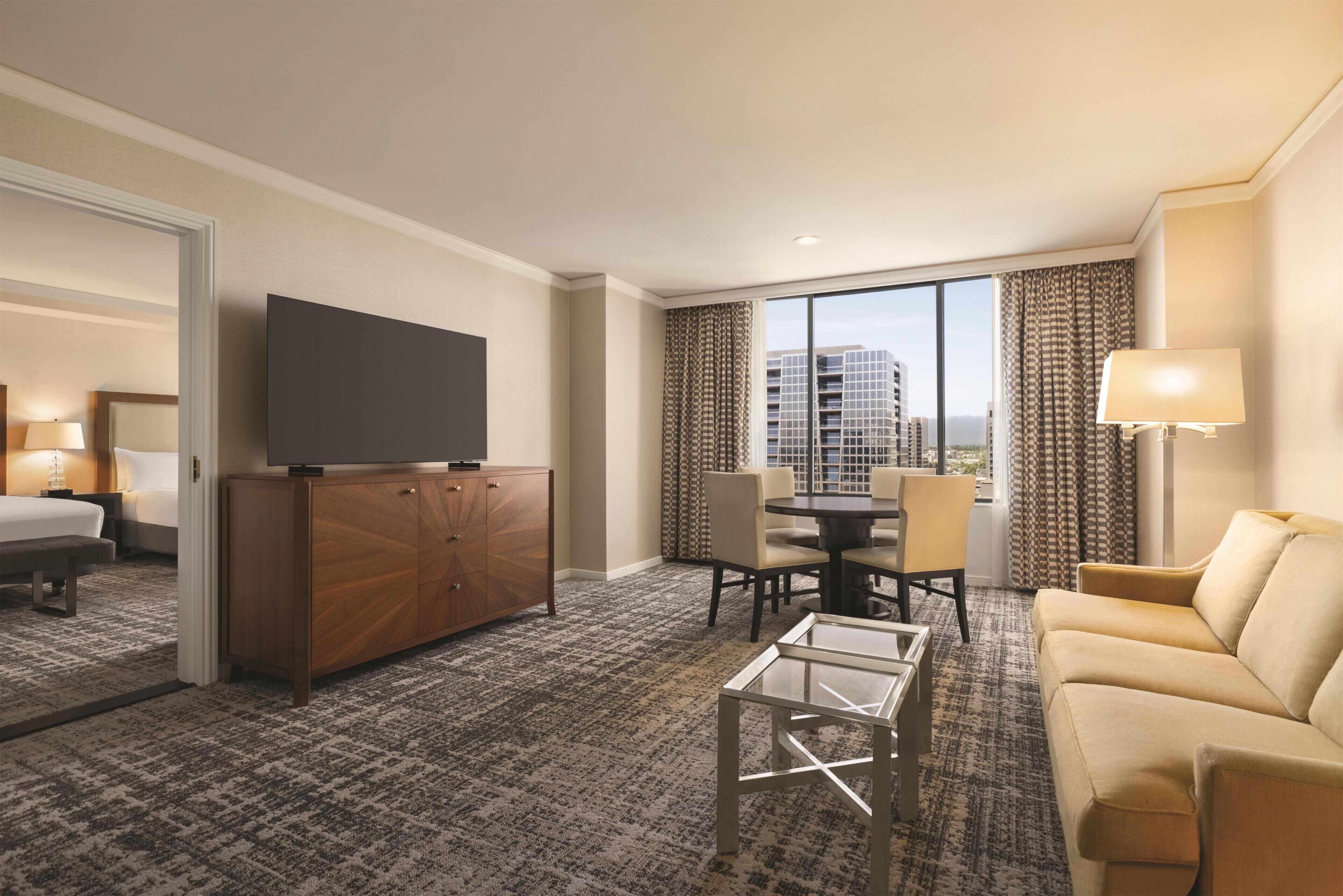 A hotel room with a bed, a TV, a dining table with chairs, a sofa, a glass coffee table, and a view of buildings through the window.