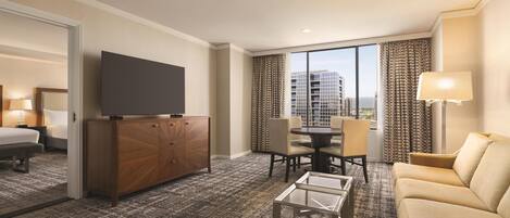 A hotel room with a bed, a TV, a dining table with chairs, a sofa, a glass coffee table, and a view of buildings through the window.