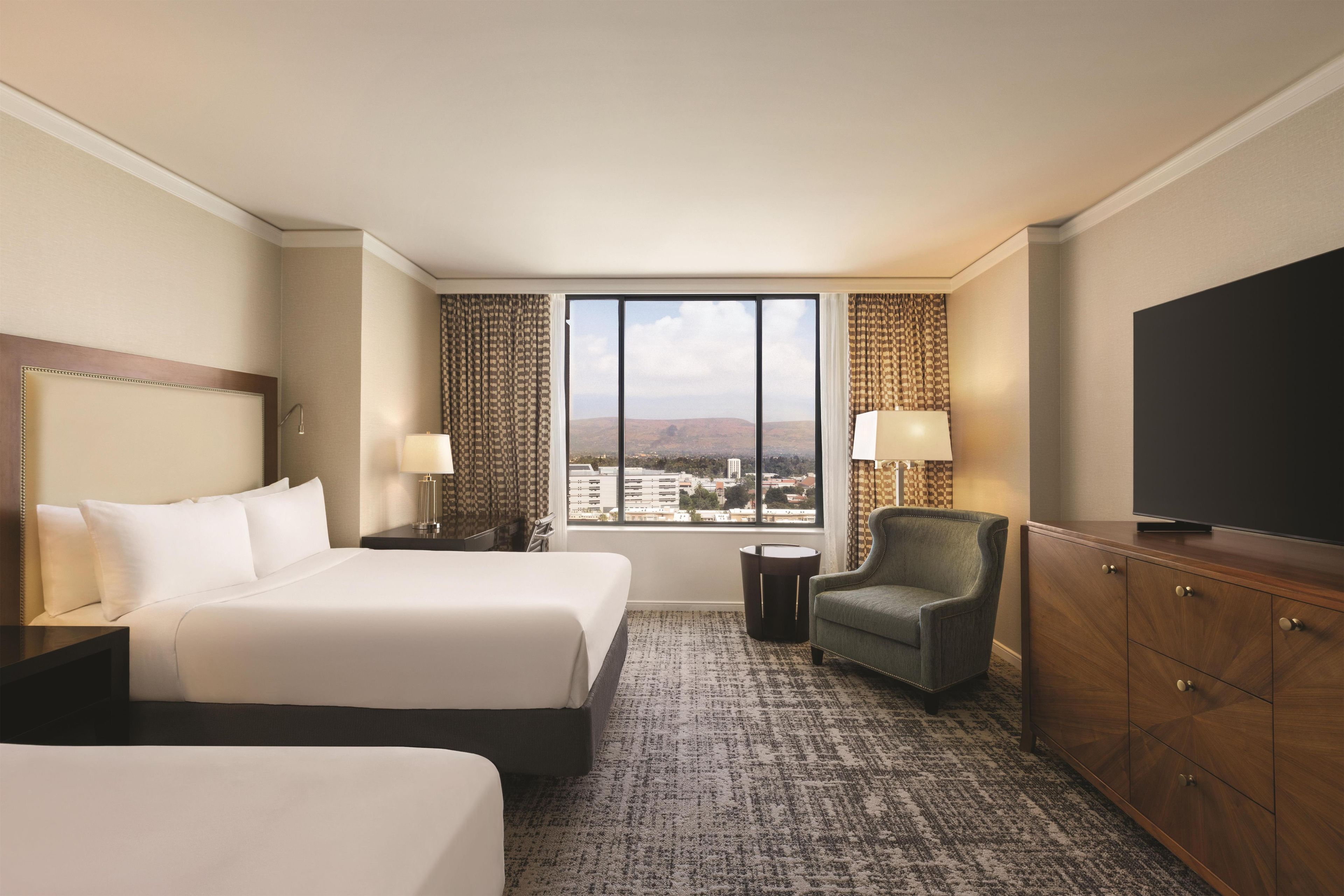 A hotel room with two beds, a TV, a chair, and a view of the city.