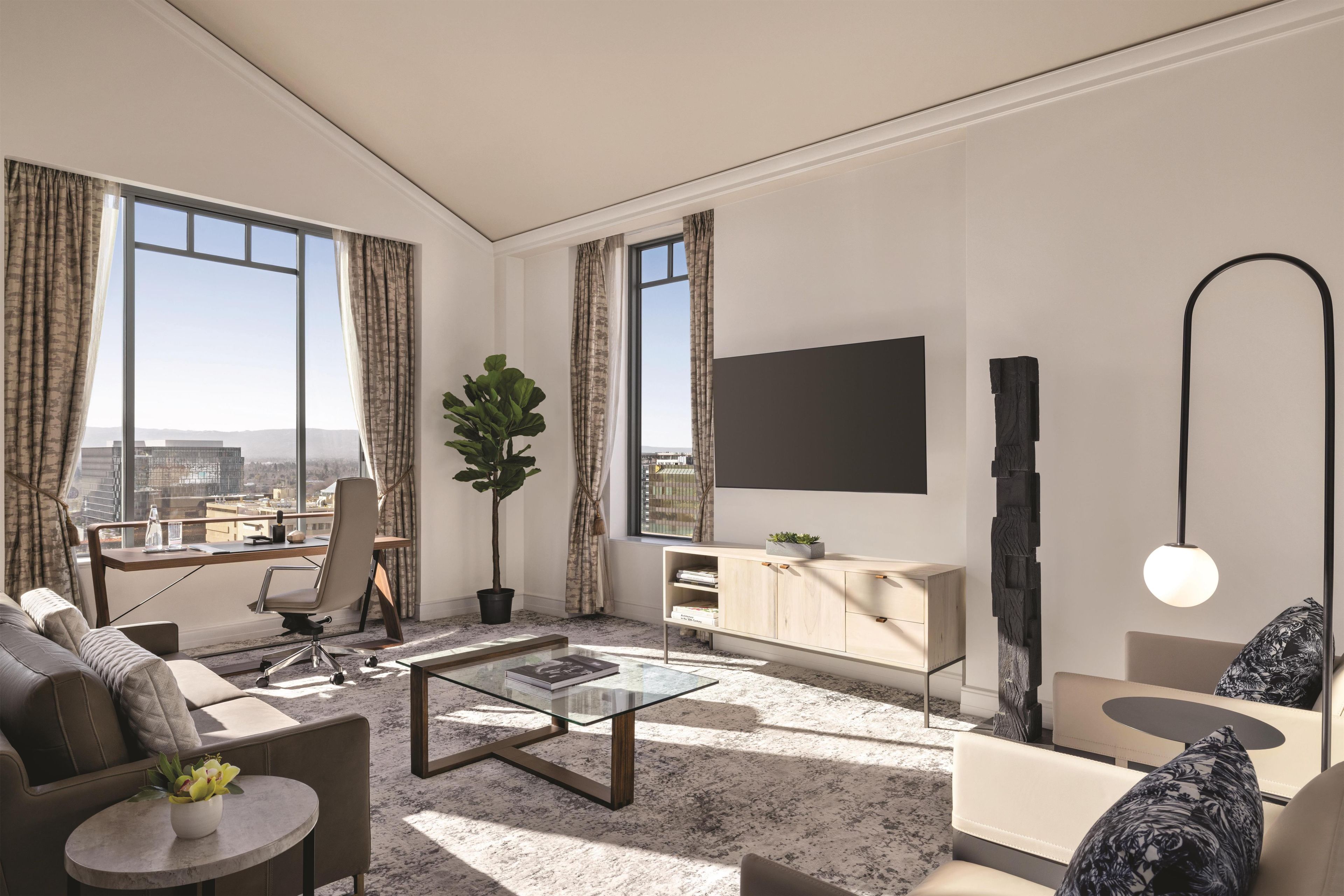 A modern living room with a sofa, coffee table, and a desk with a chair. There is a large window with curtains, a potted plant, and a flat-screen TV mounted on the wall.