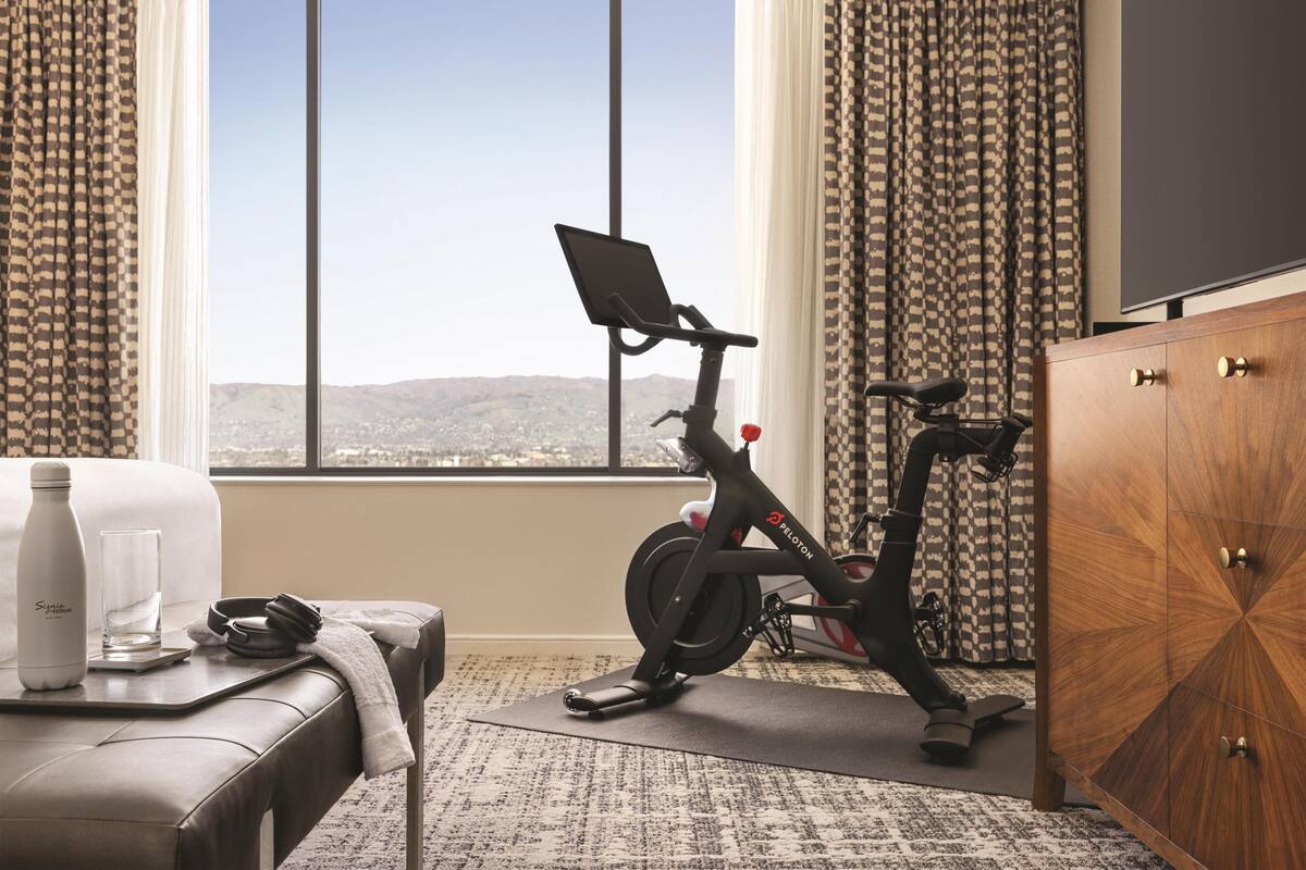 A room with a view of a cityscape, a desk with a water bottle, a glass, and a pair of headphones, and an exercise bike.