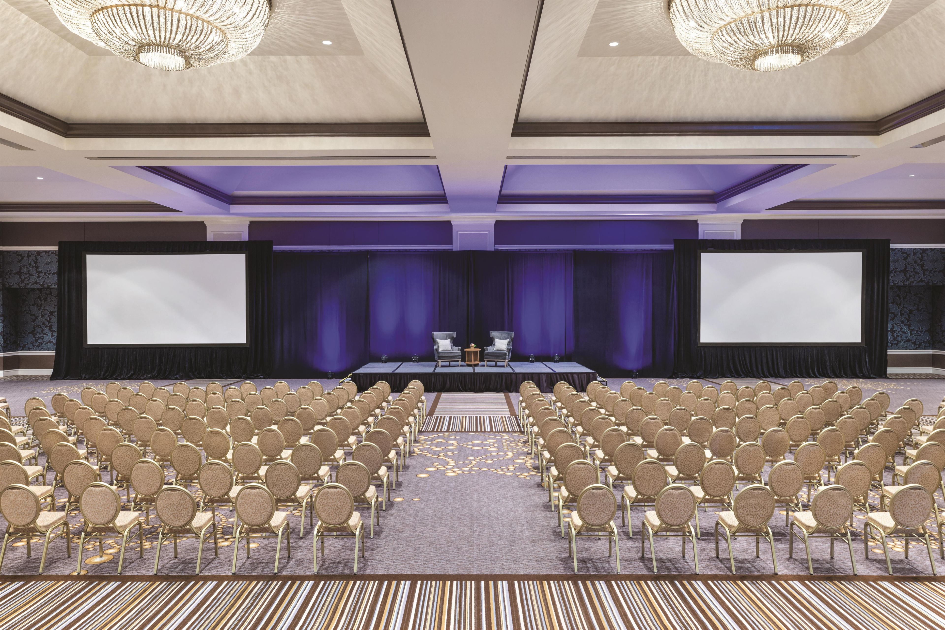A conference room with rows of chairs, a stage with a screen, and a chandelier.