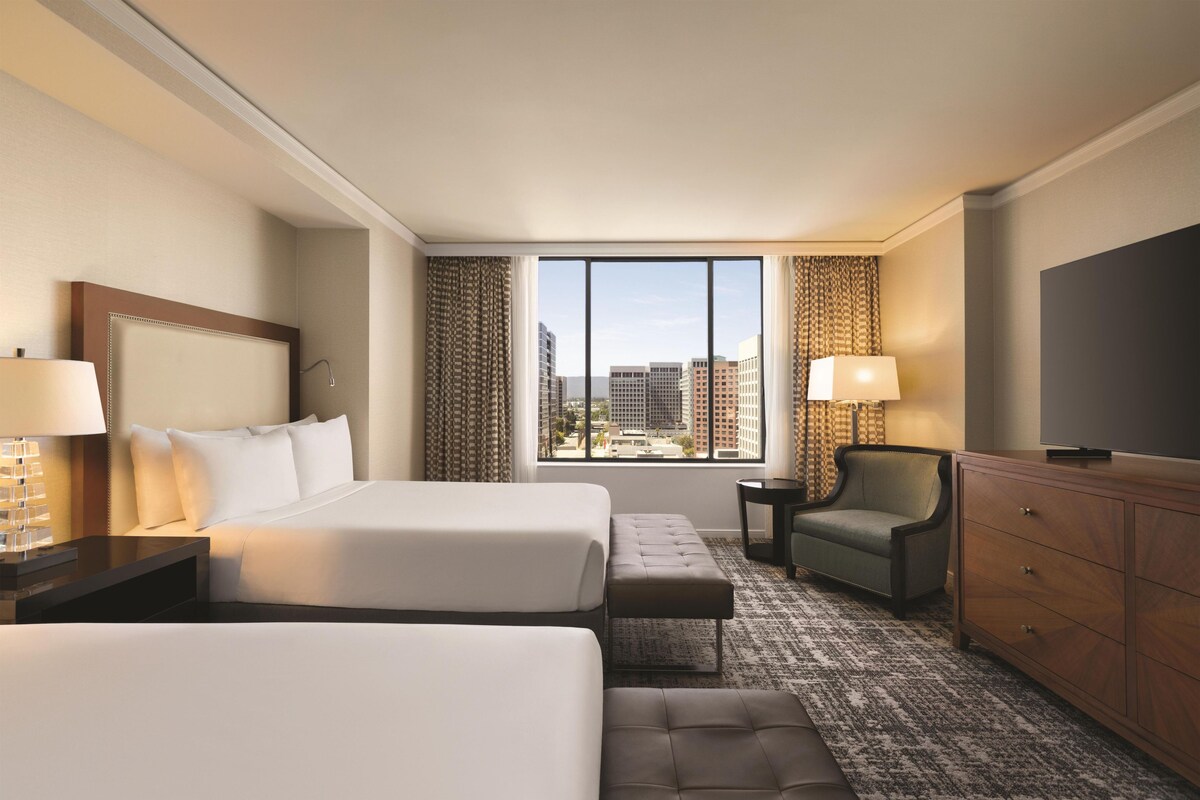 A hotel room with two beds, a sofa, a TV, a desk, and a view of the cityscape.