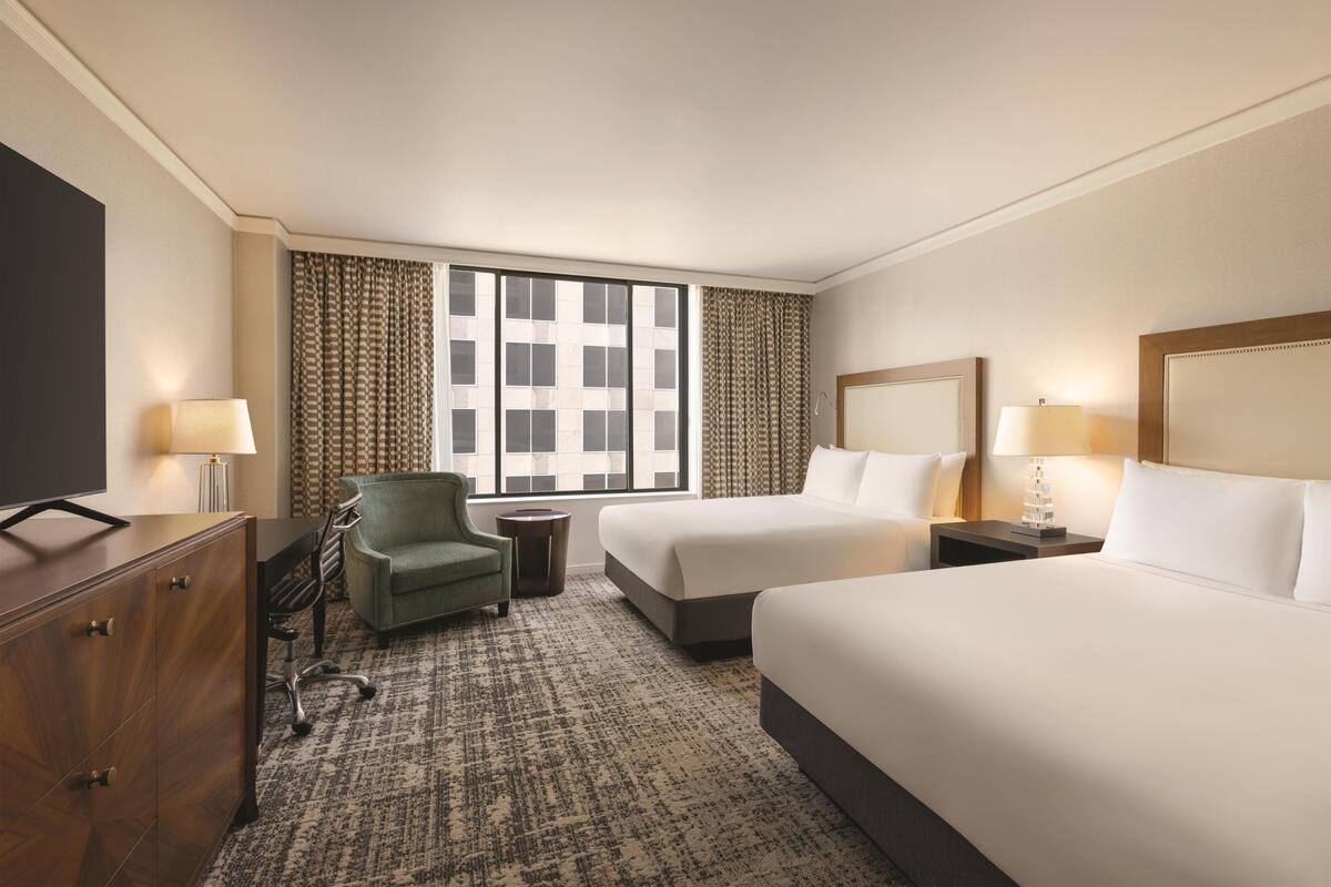A hotel room with two beds, a desk, a chair, a TV, and a window with patterned curtains.