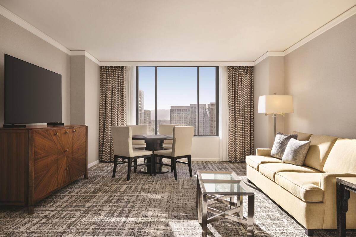 A hotel room with a large window, a sofa, a TV, a dining table, and a cabinet.