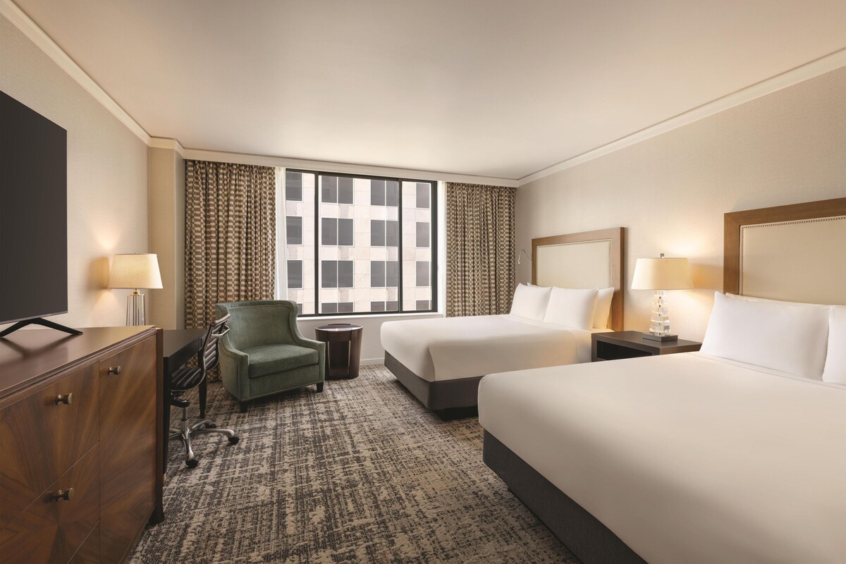 A hotel room with two beds, a desk, a chair, a TV, and a window with patterned curtains.