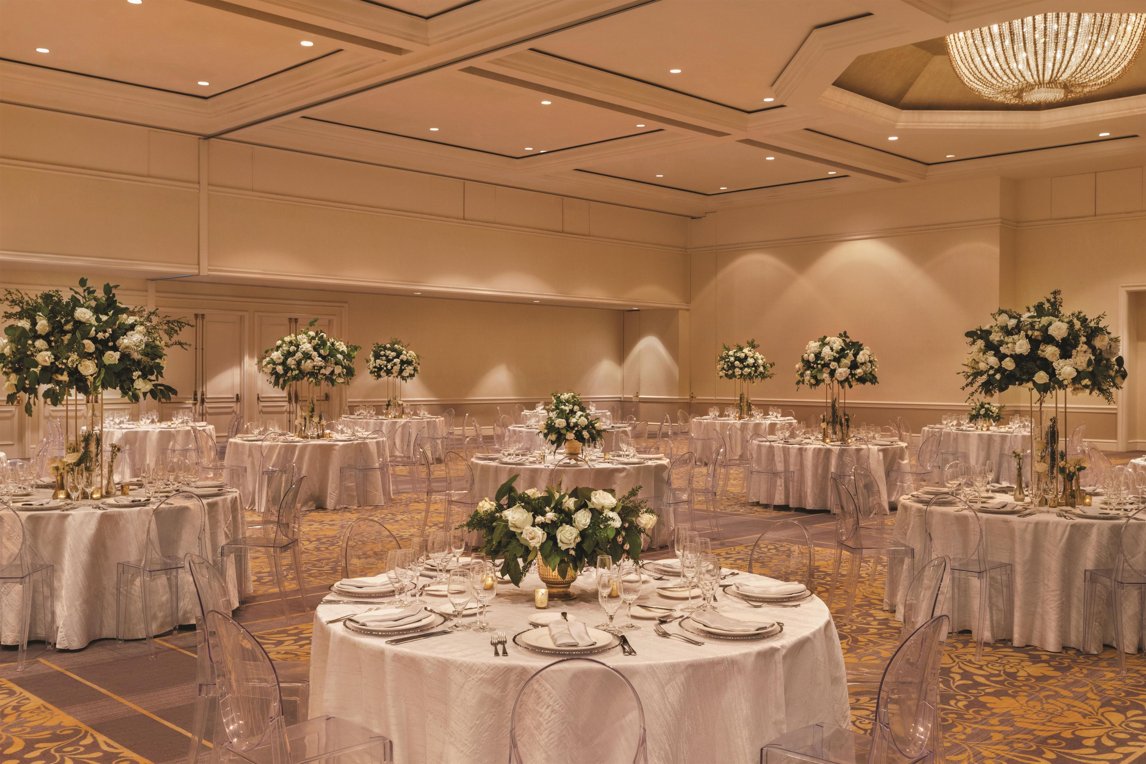 A banquet hall with round tables, clear chairs, and floral centerpieces.