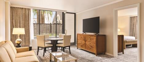 A hotel room with a sofa, a dining table with chairs, a TV on a wooden cabinet, and a balcony with a view of palm trees.